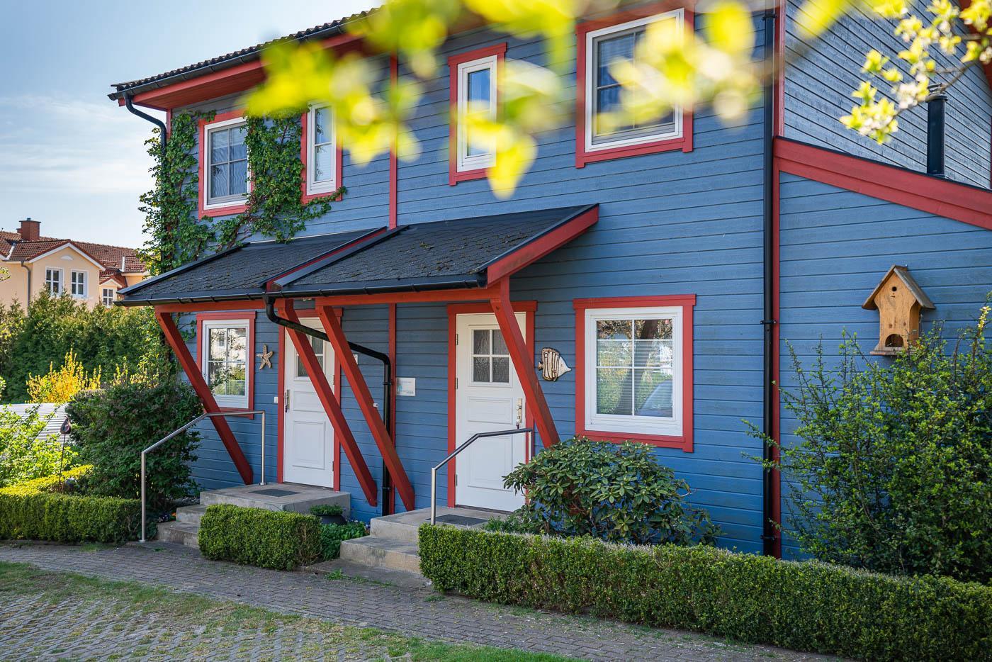 Blaues Holzhaus mit roten Fensterumrandungen. Im Vorgartenn gibt es Hecken und Sträucher.