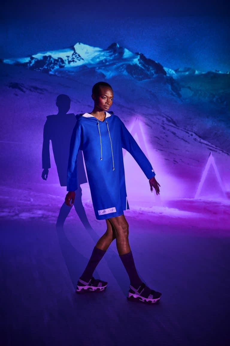 Model posiert mit einen blauen Pullover von dem Modelabel Alpin Lodge vor einer Lichtreflexion einer Gletcherlandschaft und wirft einen Schatten auf die Wand hinter sich.