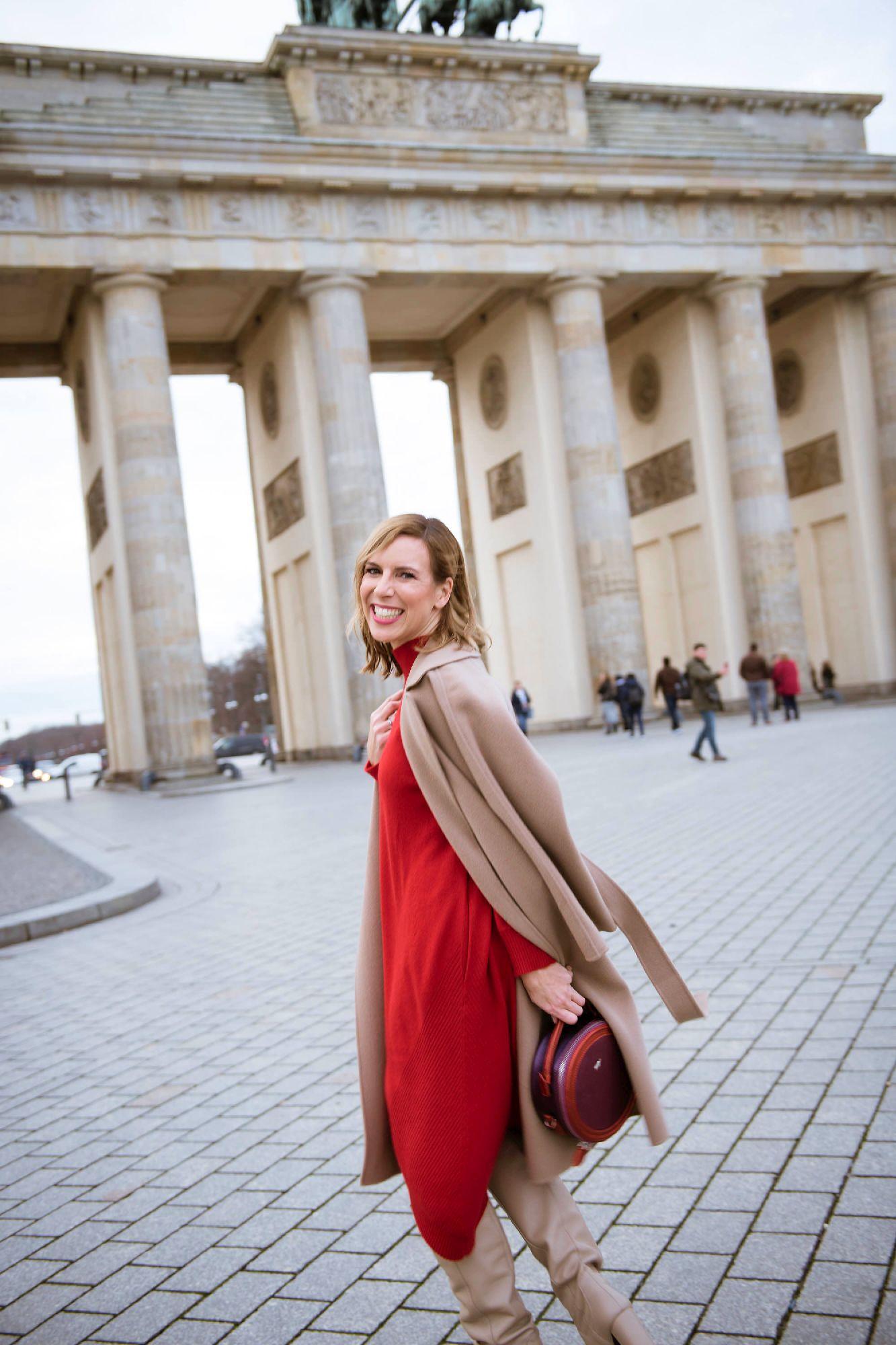 Frau mit rotem Kleid und beiger Jacke sowie Stiefeln vom dem Modelabel Edelziege, dreht sich lachend in die Kamera, während sich das Brandenburger Tor im Hintergrund befindet.