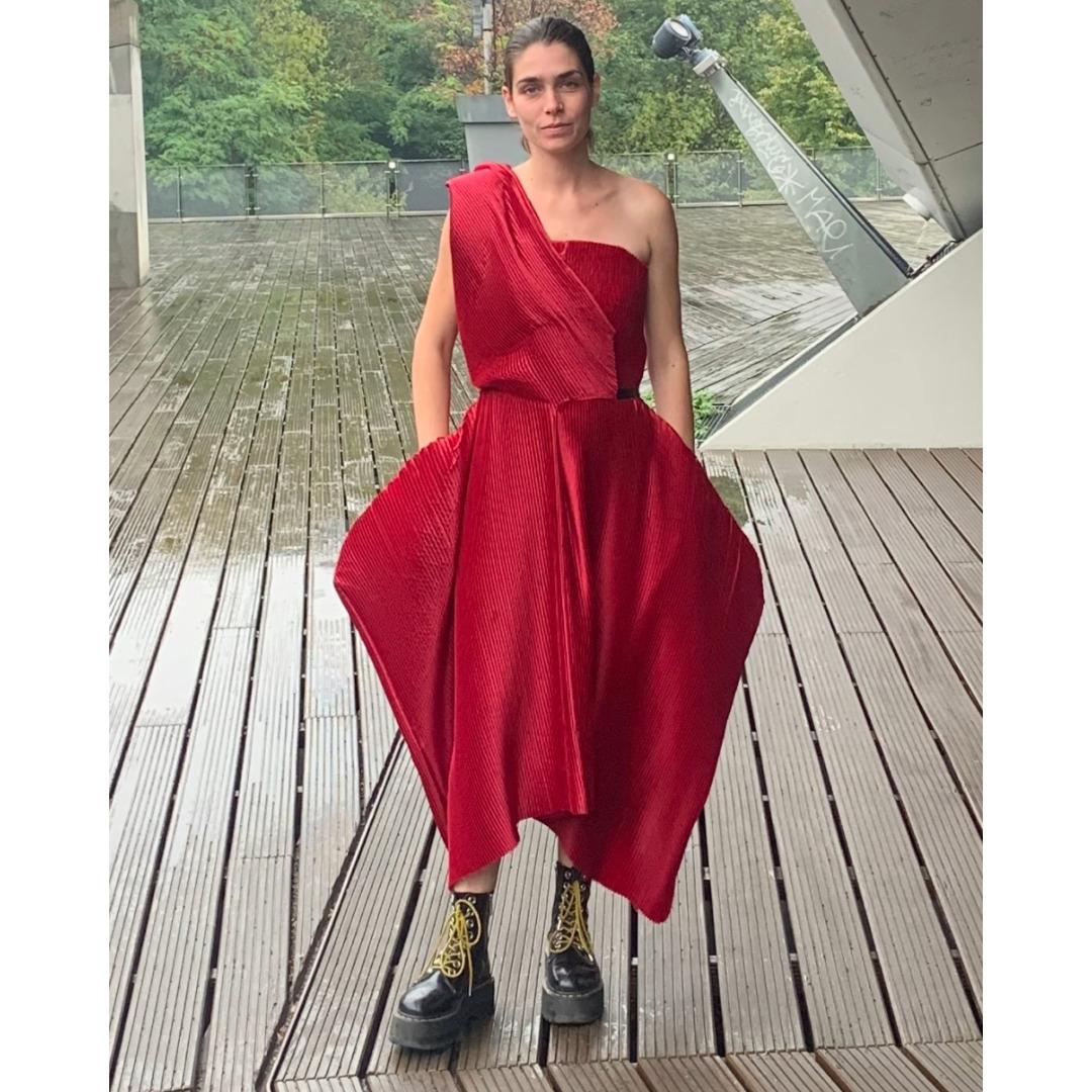 Frau mit braunen zusammengebundenen Haaren trägt auf einer großen verregneten Terrasse ein rotes Kleid vom Modelabel Timmhoth. Dazu trägt Sie schwarze Stiefel mit gelben Schnürsenkeln