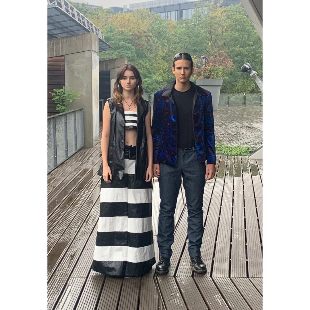 Männliches und weibliches Model stehen mit neutralen Gesichtsausdruck auf einer verregneten Terrasse mit Kleidung vom Modelabel Timmhoth. Die Frau trägt einen langen Rock im Zebramuster, sowie ein Top in der selben Farbei, sowie eine schwarze Weste darüber. Der Mann trägt Stiefel mit einer Anzugsose, sowie ein schwarzes T-Schirt mit einem schwarzen Sakko, welches blaue edle Akzente aufweist.