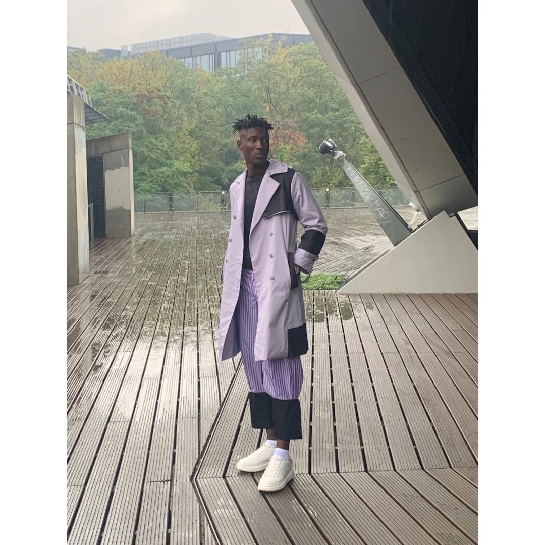 Mann steht auf einer verregneten Terrasse mit weißen Sneakern und lavendelfarbender Hose. Diese hat feine schwarze Streifen und dazu trägt er ein scharzes T-Shirt, sowie einen langen lavendelfarbenden Mantel von dem Modelabel Timmhoth