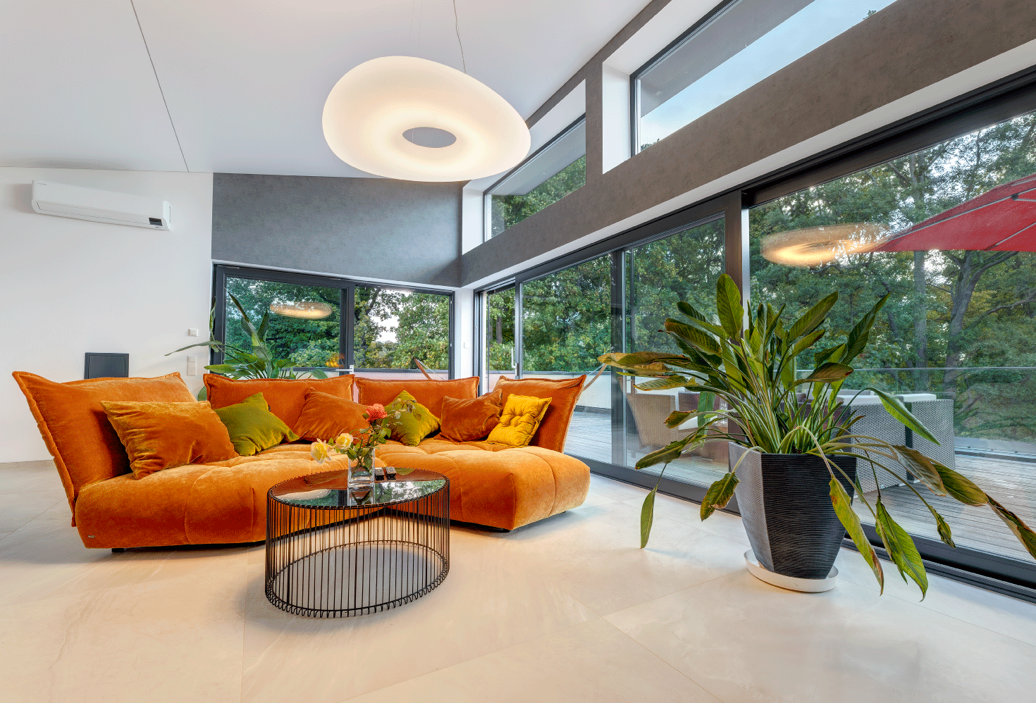 Innenansicht des Penthouses mit einem gemütlichen, modernen Sofa in Orange, welches vor einem Panoramafenster steht. Die Ausicht aus dem Panoramafenster zeigt große, grüne Laubbäume 