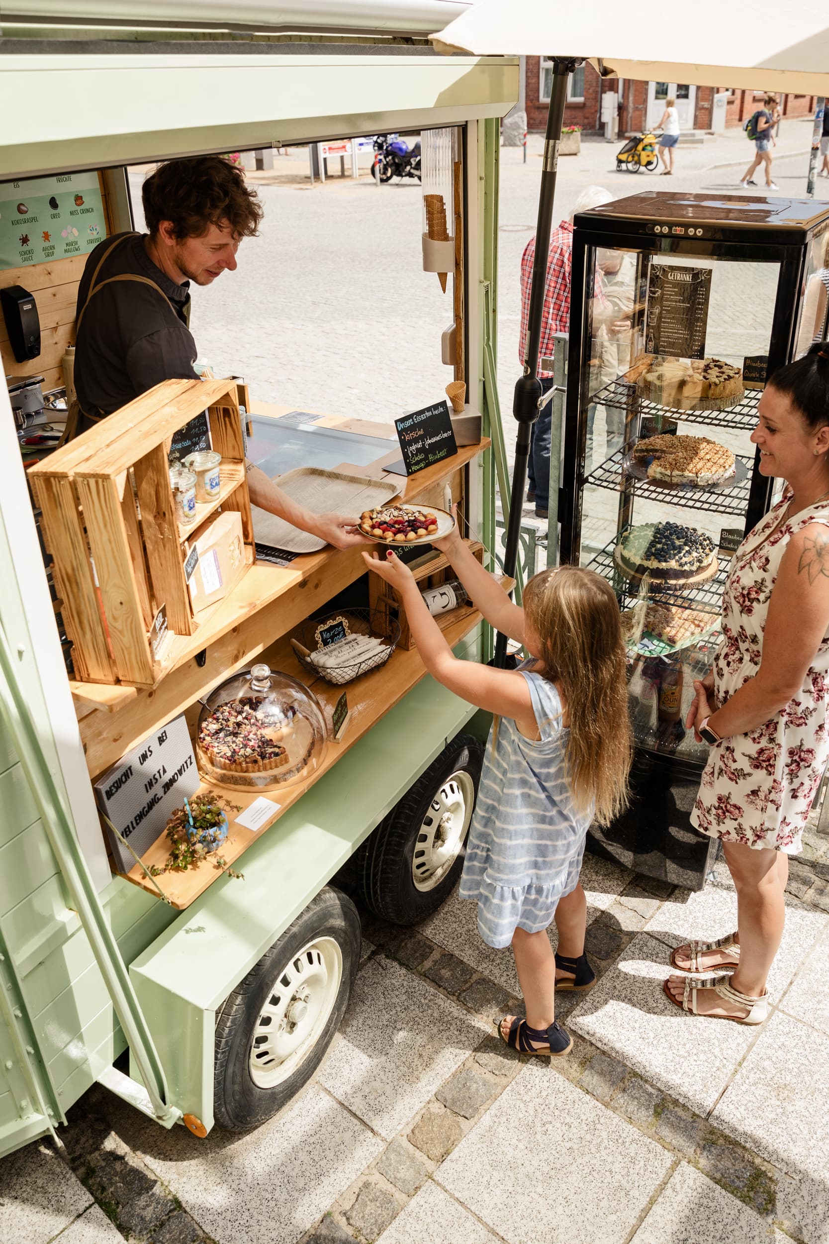 Ein männlicher Verkäufer verkauft aus einem Foodtruck kuchen. Ein Kind bekommt Kuchen überreicht