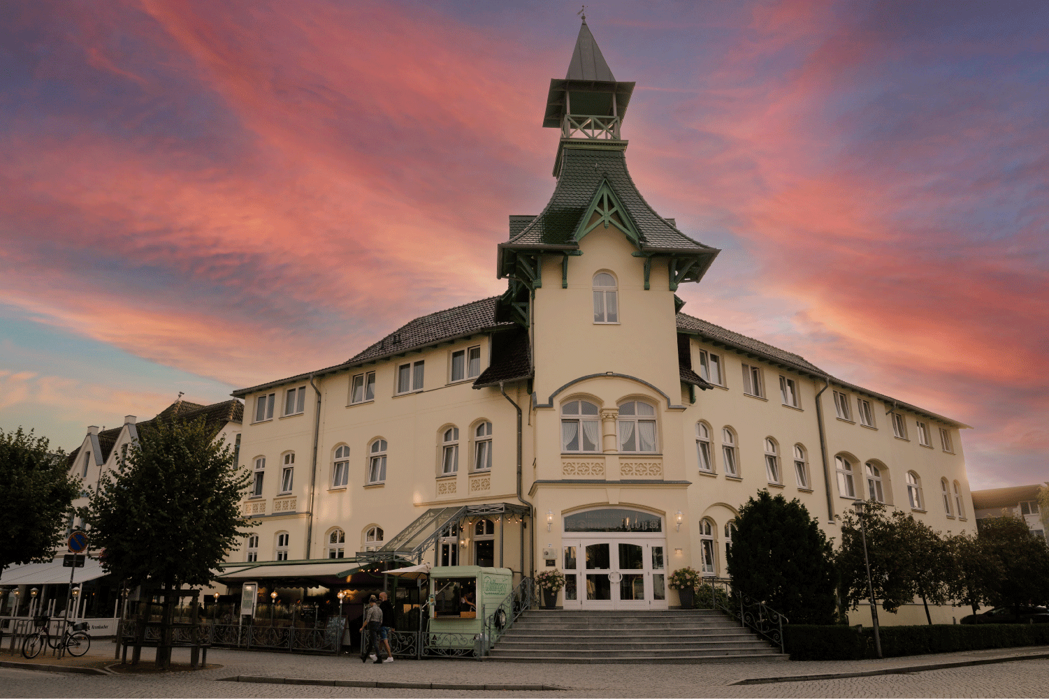 Frontalansicht eines im Bäderhaus-Stil gebauten Hauses mit einem Türmchen. Im Hintergrund sind lila-blaue Wolken, die durch die Abendsonne entstehen