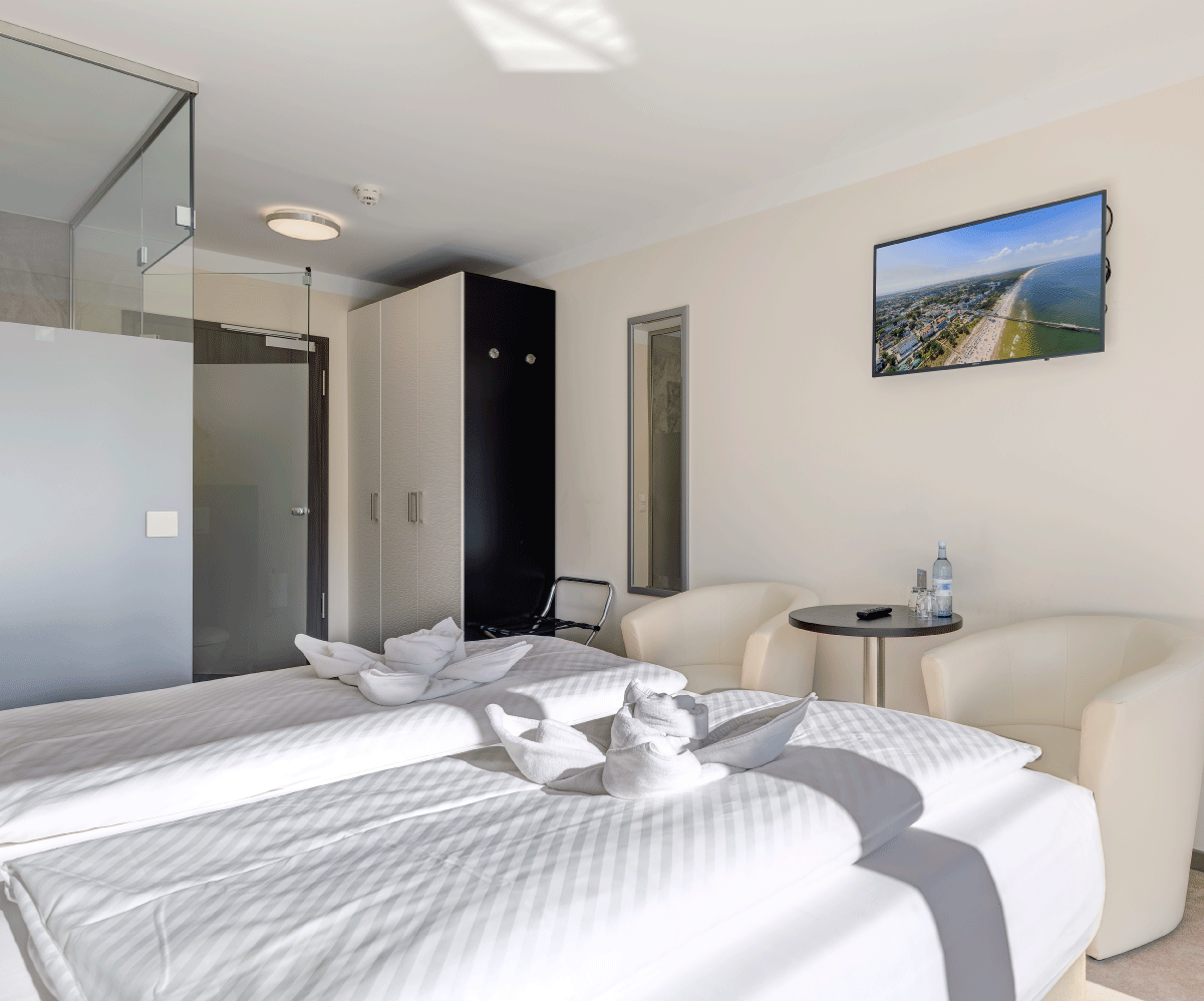 gemütlich eingerichtetes Zimmer mit Doppelbett, zwei weißen Ledersesseln und einem Fernseher an der Wand 