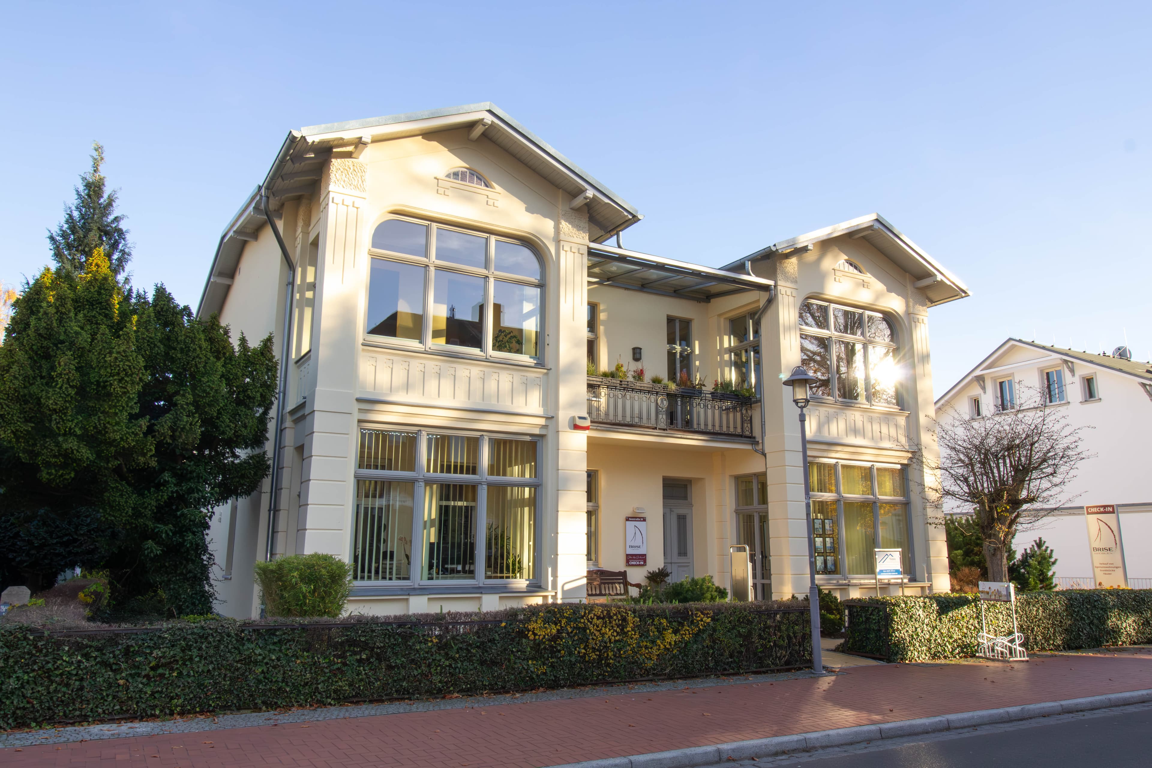 frontale Aussenansicht der Villa Brise. Ein Cremefarbenes großes Haus mit lichtdurchfluteten Fenstern einem begrüntem Vorgarten und einem rot gepflasterten Gehweg davor 