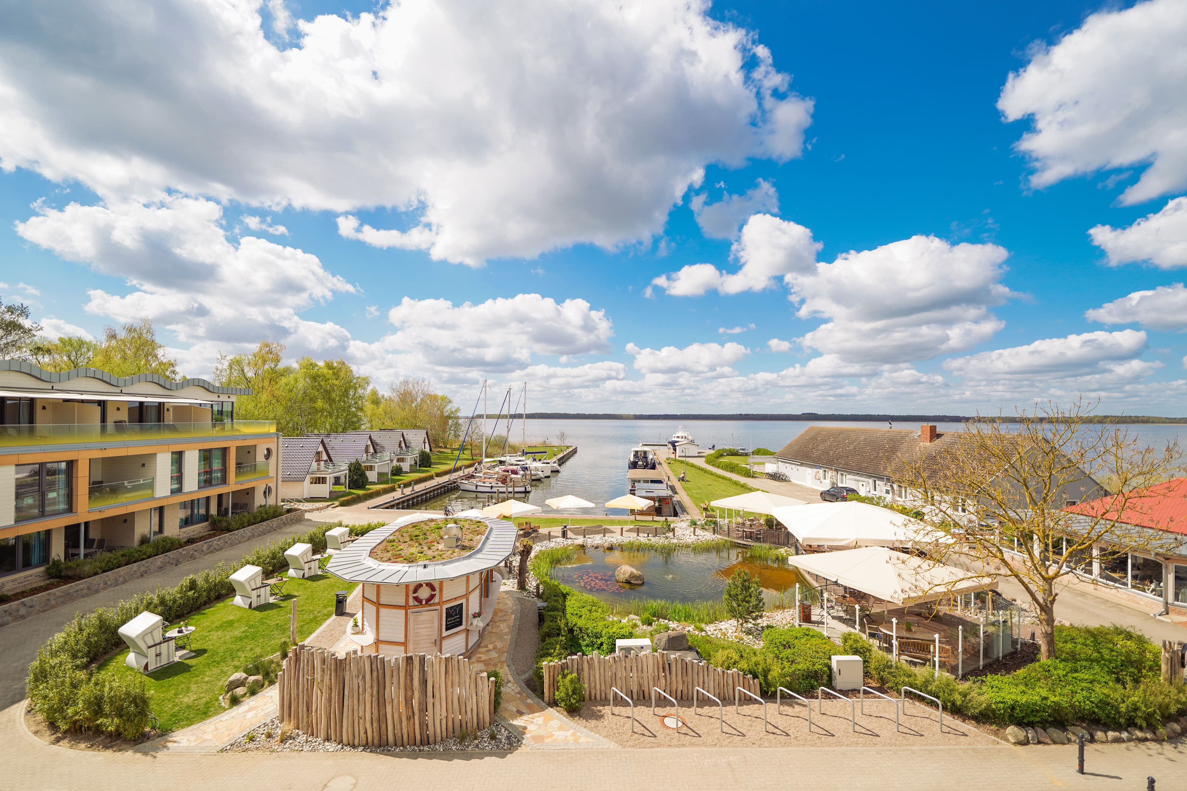 blauer Himmel mit weißen Wolken, begrüntes kleines Hafenbecken, drumherum sind Appartements und ein Fischrestaurant sowie Fischverkauf
