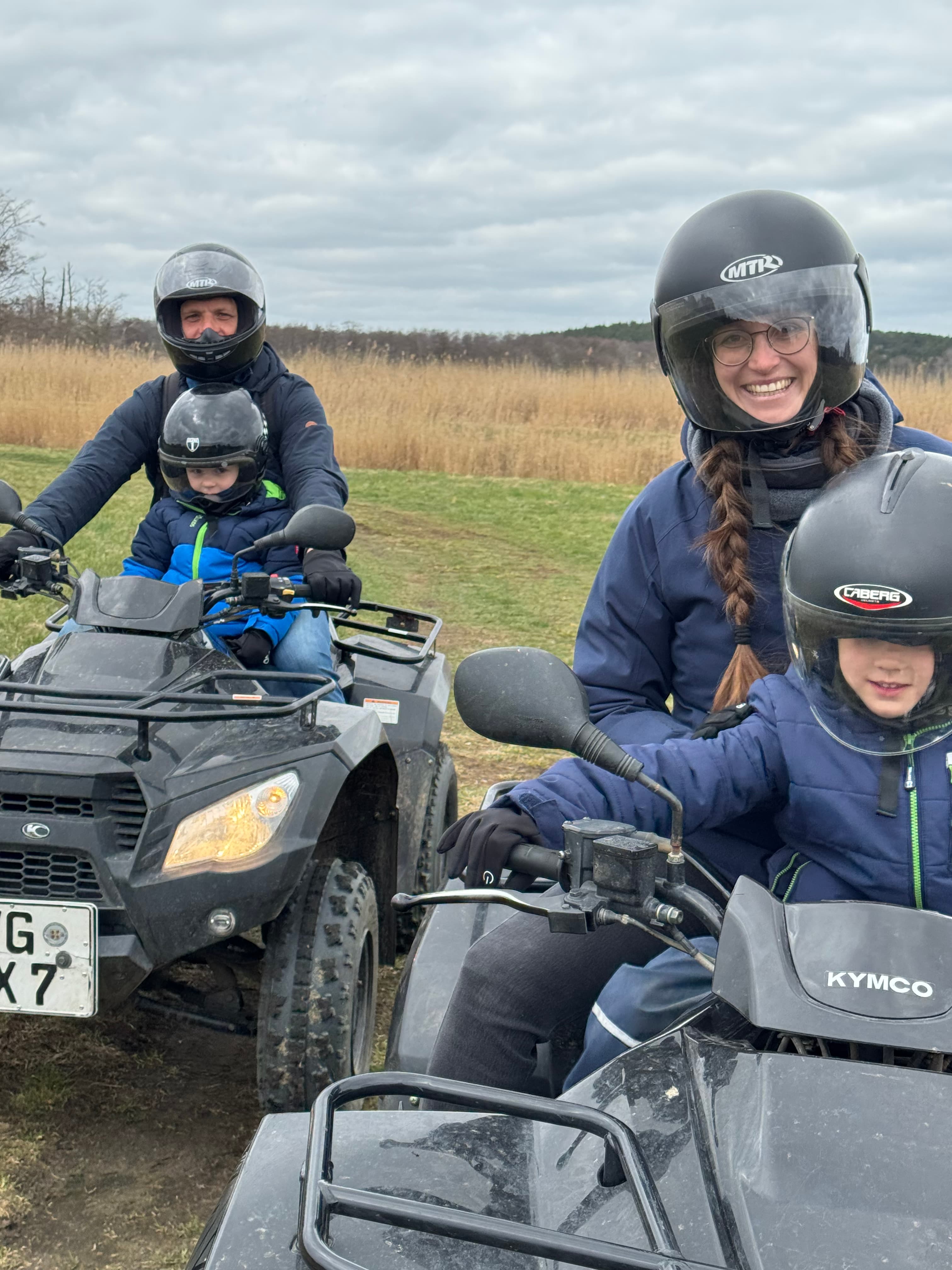 Eine 4-köpfige Familie macht eine Quad-Fahrt. Mama und Papa sitzen jeweils auf einem Quad und vor ihnen hat jeder ein Kind. Alle lächeln.