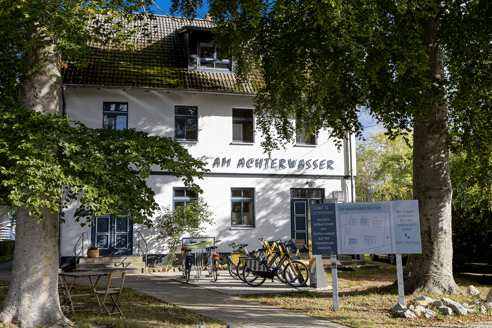 Eingangsgebäude der Ferienwohnanlage, mit großen Bäumen im Vordergrund, Begüßungsschild und Fahrrad Verleihstation