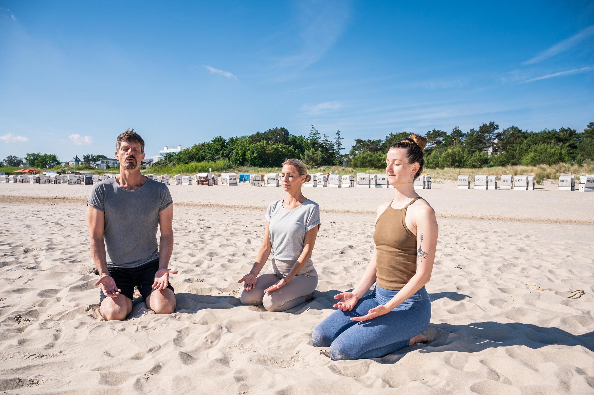 Drei Personen beim Meditieren beim Yoga am Strand.