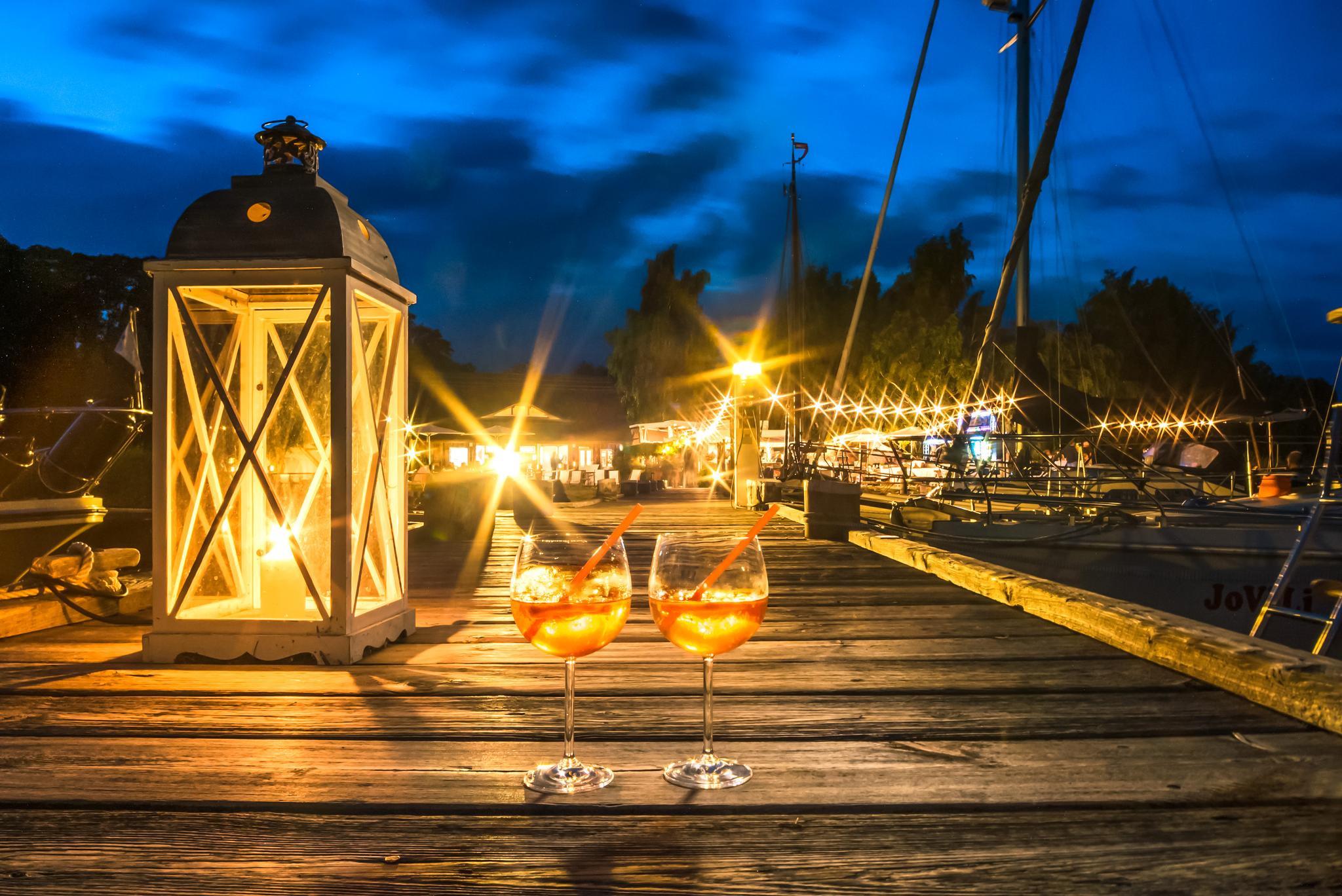Weingläser und Laterne auf einem Steg. Im Hintergrund beleuchteter Naturhafen..