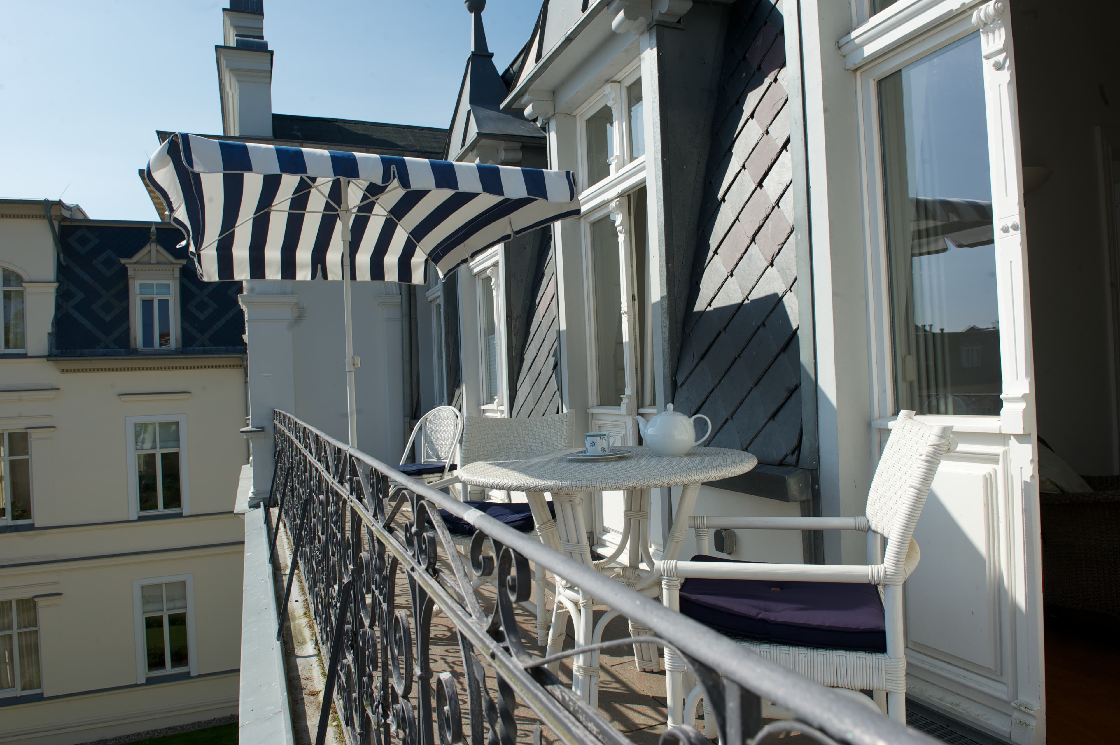 Weißer Balkon mit weißen Sitzmöbeln und einem weiß-blau gestreiften Schirm.