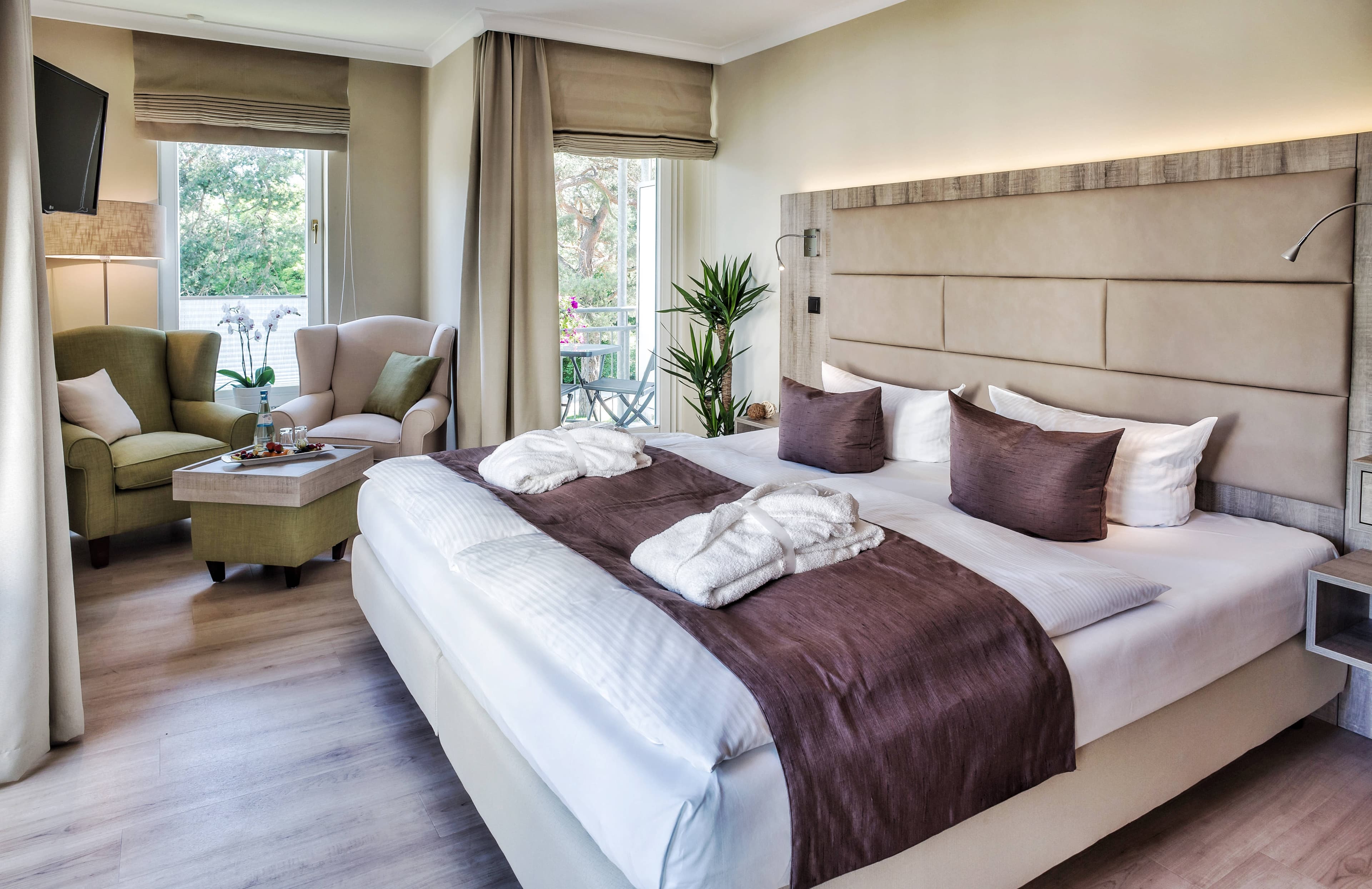 Doppelbett und Sessel im Hotelzimmer. Blick geradaus auf den Garten des Hotels.