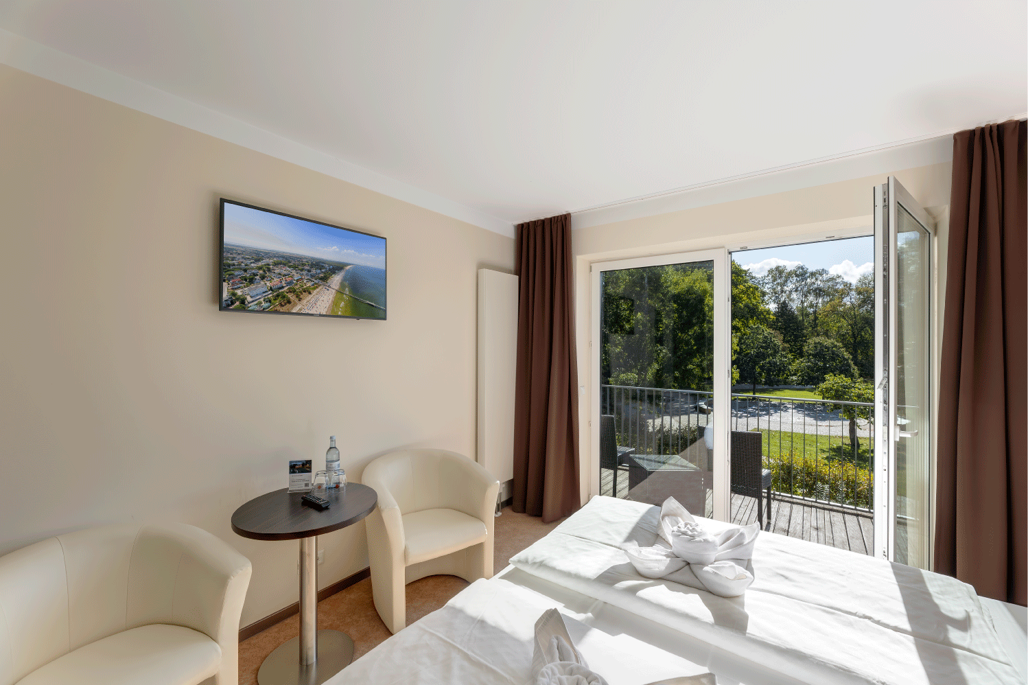 gemütlich eingerichtetes Zimmer mit Doppelbett, zwei weißen Ledersesseln und einem Panoramafenster mit Blick auf den grünen Park